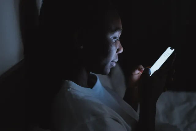 A woman sitting in a dark bedroom at night, her face lit by the glow of her phone screen