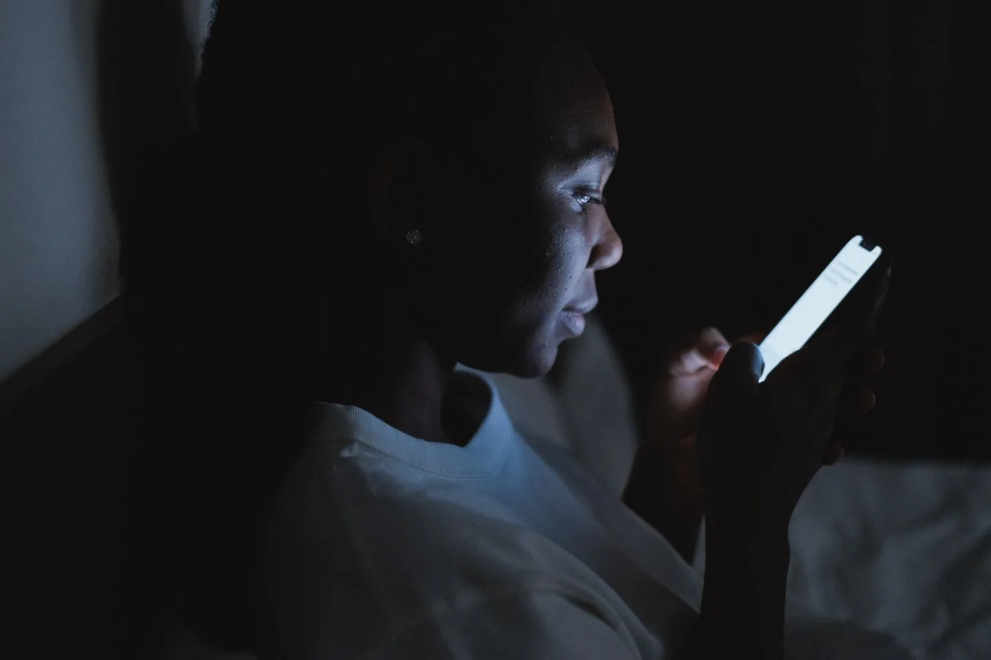 A woman sitting in a dark bedroom at night, her face lit by the glow of her phone screen