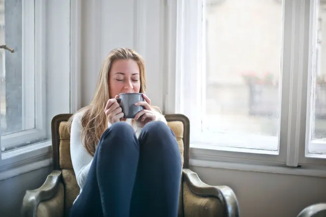Woman wrapped in a sweater holding a warm mug in an armchair by a window