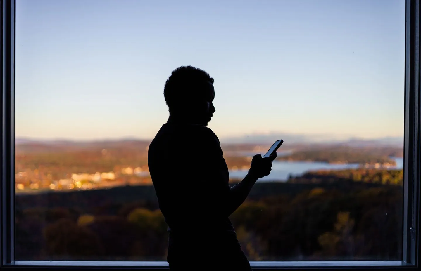 A person silhouetted by a window at twilight, looking down at their phone
