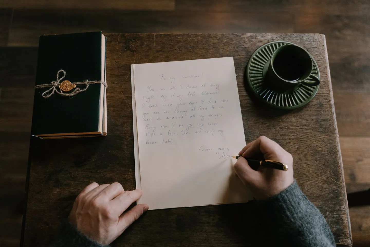 A person handwriting a letter at a wooden table beside a journal and a cup of coffee