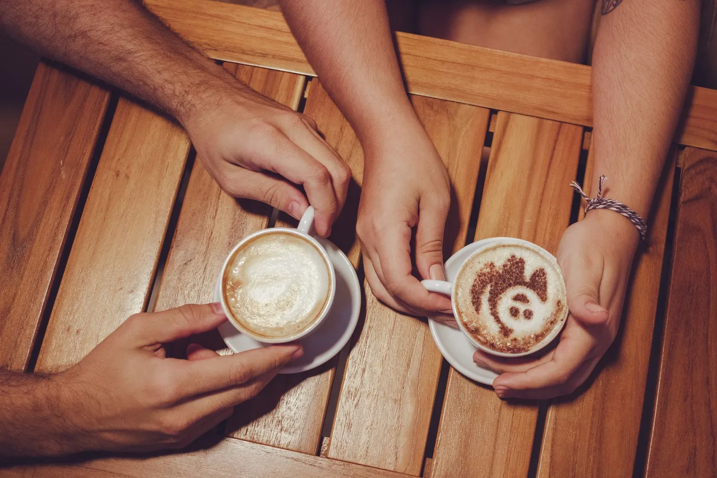 Two people holding coffee cups with latte art across a wooden table