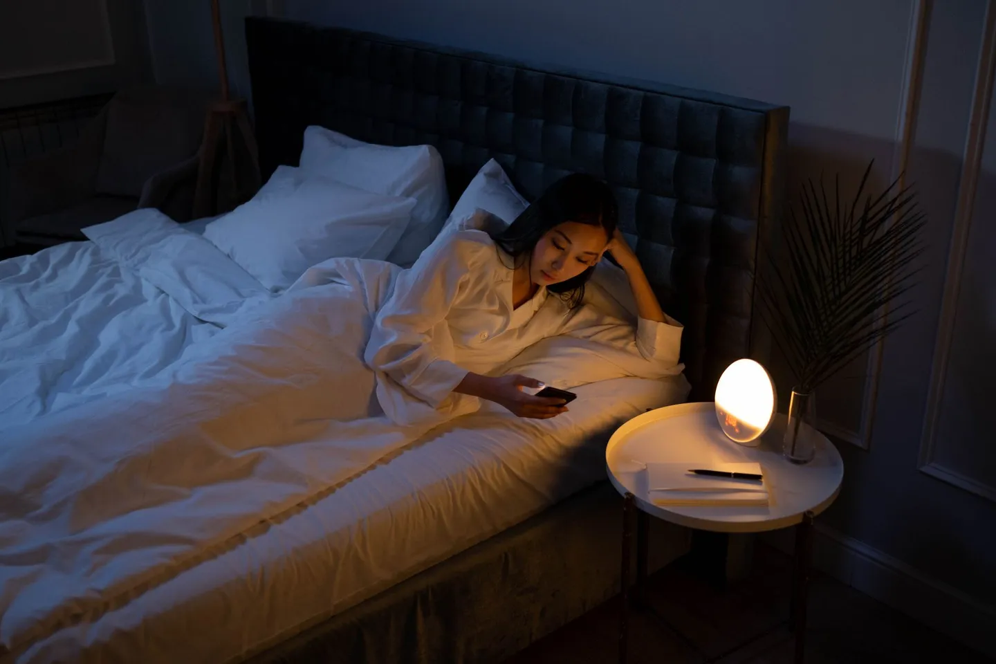 A woman lying awake in a dark bedroom at night, looking at her phone beside a glowing lamp