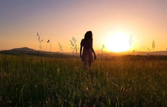 A woman standing in a green field facing a vibrant sunset sky
