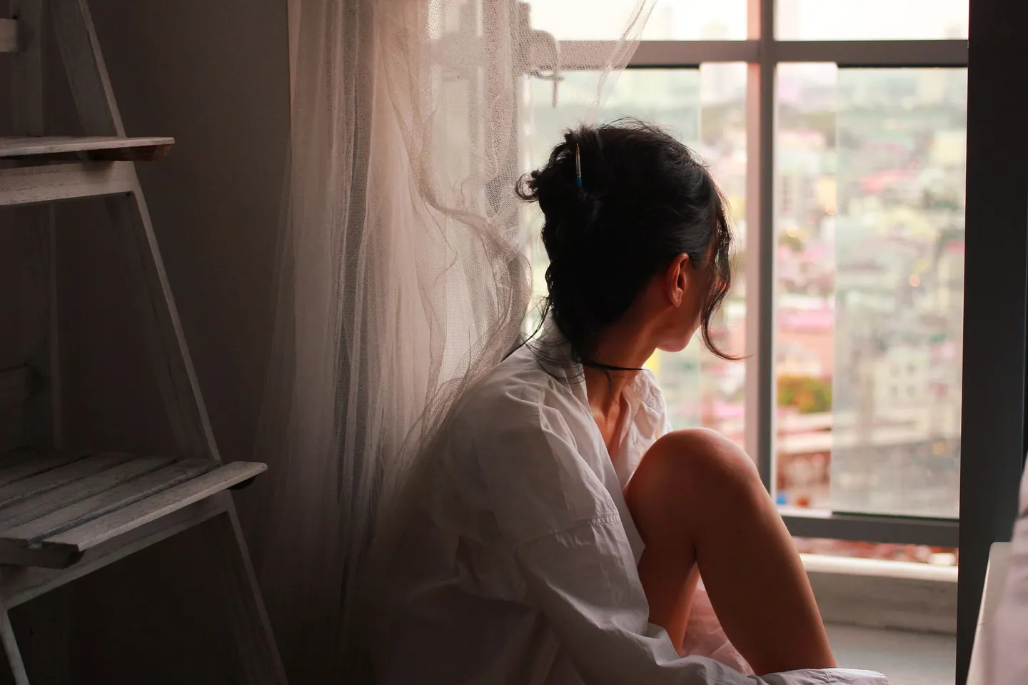 A woman sitting by a large window at dusk, gazing out at the city below